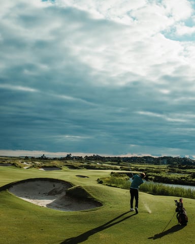 Golfer mid-swing on a coastal links course with sand bunkers and dramatic sky