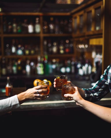 Two people clinking whiskey glasses at a dark bar with bottles behind