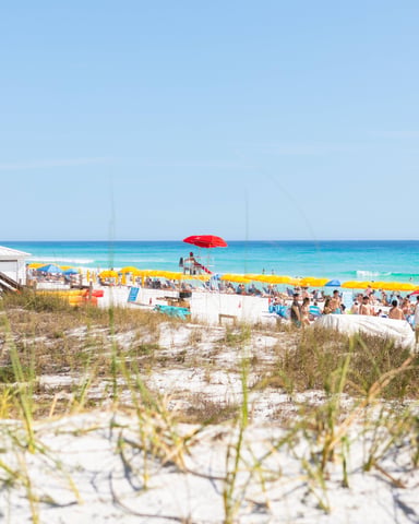 Sandy beach with dune grass, crowded with colorful umbrellas and beachgoers, turquoise water and clear sky