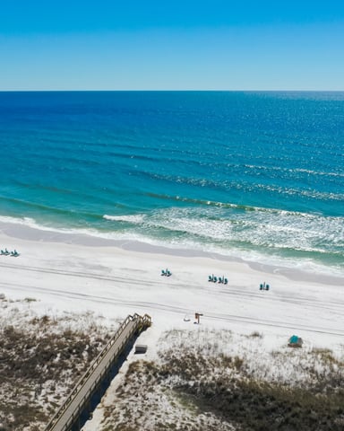 Aerial view of sandy beach with blue umbrellas, wooden boardwalk, dune vegetation, and turquoise ocean with breaking waves