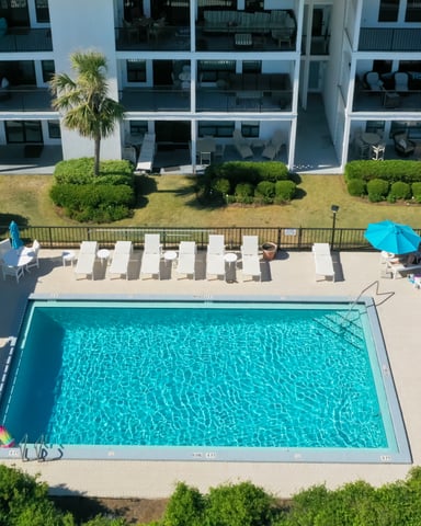 Aerial view of resort pool with turquoise water, lounge chairs, blue umbrellas, and modern white building with balconies