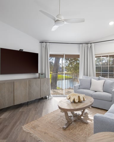 Bright living room with gray sofas, wall-mounted TV, wooden coffee table, and glass doors opening to a balcony overlooking green landscape.