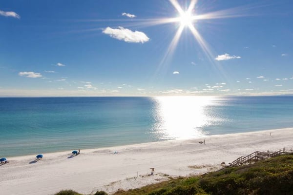 Sunny beach with white sand, turquoise water, blue umbrellas, and wooden beach access stairs