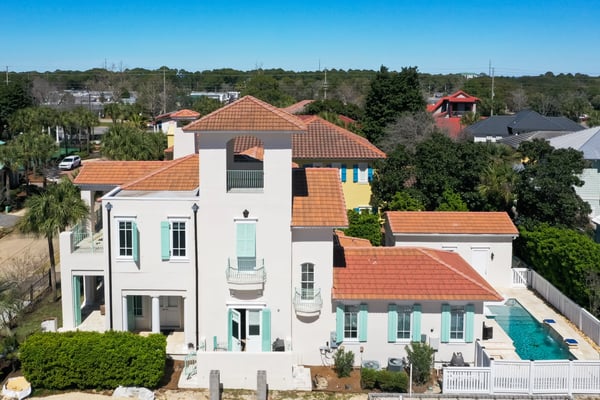 Aerial view of white Mediterranean-style home with terracotta tile roof, turquoise shutters, and backyard swimming pool surrounded by white fence