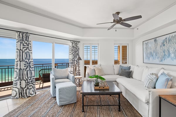 Bright beachfront living room with ocean view, white sectional sofa, patterned curtains, and balcony access