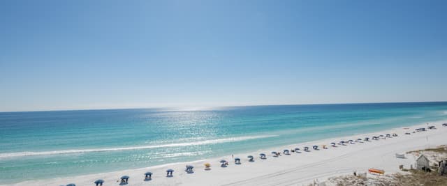 Wide sandy beach with turquoise water, rows of blue umbrellas, and bright sunny sky