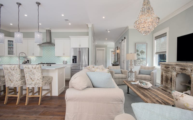 Open-plan kitchen and living room with white cabinetry, turquoise backsplash, stainless steel appliances, beige upholstered seating, crystal chandelier, and