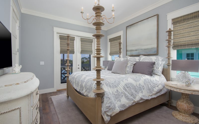 Master bedroom with four-poster bed, brass chandelier, blue-gray walls, and French doors opening to a deck with ocean view