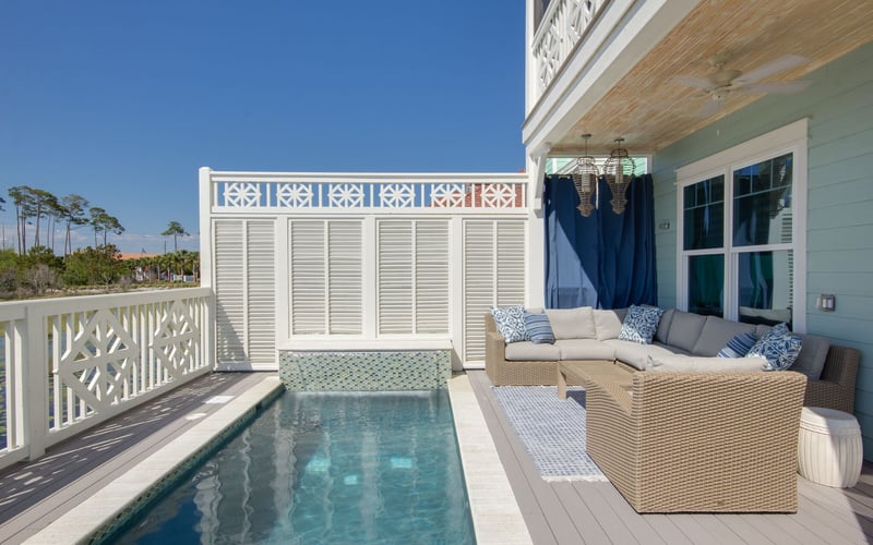 Coastal deck with small pool, wicker seating, blue curtains, and white railing overlooking trees and clear sky