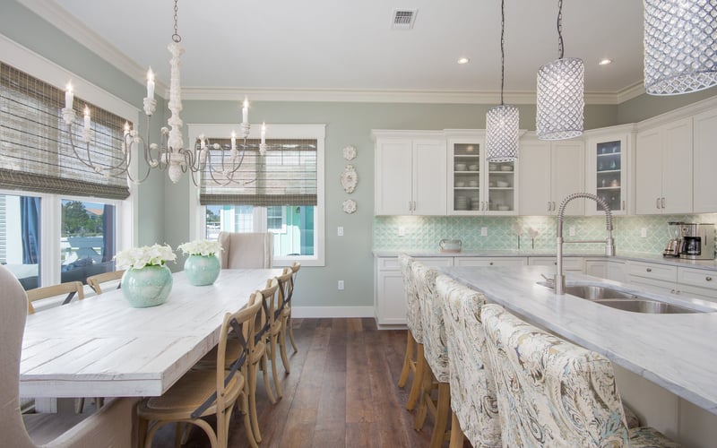 Open-plan kitchen and dining area with white cabinetry, marble countertops, mint-green walls, and water views through large windows.