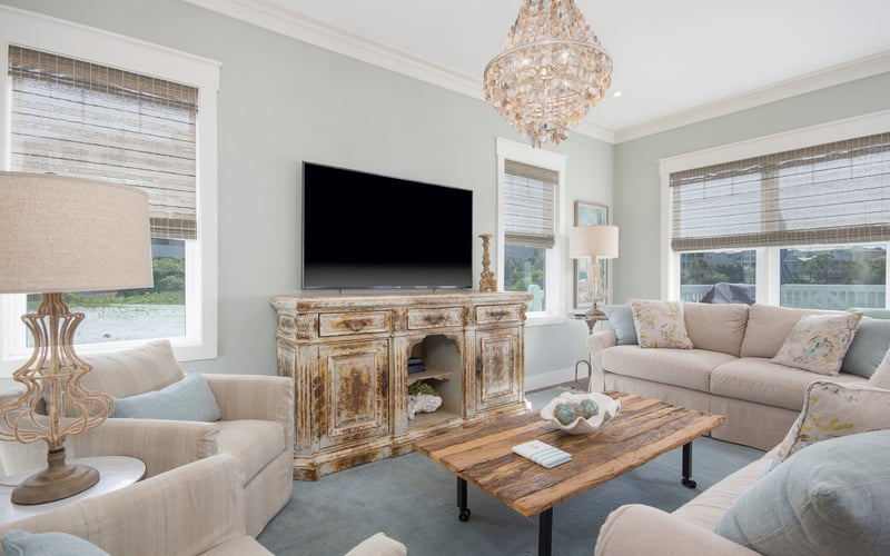 Bright living room with cream upholstered seating, distressed wood TV console, crystal chandelier, and large windows with woven shades.